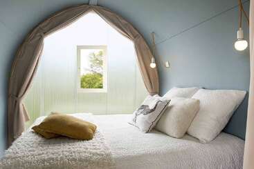 Cette chambre confortable et minimaliste est dotée d'une fenêtre ronde, d'une literie blanche douce, de coussins décoratifs, de suspensions lumineuses, de murs bleus et d'une lumière naturelle qui créent une atmosphère sereine et accueillante.