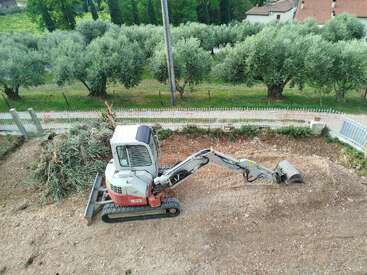 Ein kleiner Bagger arbeitet auf einem unbebauten Grundstück und räumt und ebnet den Boden in der Nähe eines Haufens von Ästen. In der Nähe sind Olivenbäume und ein Zaun zu sehen.