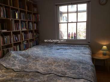 The image depicts a bedroom with a bed, bookshelf, and window, featuring a blue and white patterned comforter and a wooden nightstand with a lamp.