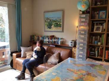 The image depicts a woman sitting on a brown leather couch in a cozy room, cradling a small white dog. She is dressed in a dark blue outfit.
