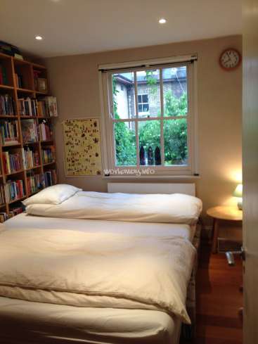The image depicts a cozy bedroom with a bed, bookshelf, and window, featuring a white duvet and pillows, and a clock on the wall.