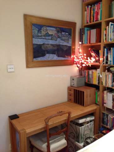 The image depicts a cozy corner of a room, featuring a wooden desk, chair, bookshelf, and painting, with a small tree adorned with lights adding a touch of warmth.