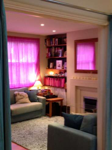 The image depicts a cozy living room with a fireplace, bookshelf, and couches, featuring pink curtains and a grey rug, with a watermark reading "worldway.info".
