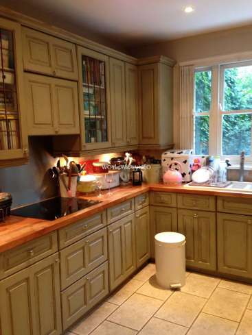 The image depicts a kitchen with sage green cabinets and a butcher block countertop, featuring a sink, stove, and various appliances.