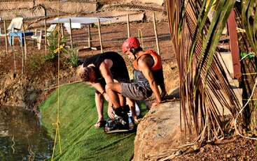 Two men prepare for water sports by a lake; one assists the other with boots and gear. Both display concentration and teamwork under the afternoon sun.