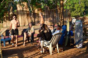 A group of people relax outdoors on plastic chairs, enjoying drinks and conversation. Surfboards and trees surround them, suggesting a casual beach or surfside gathering.