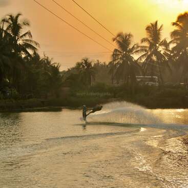 A person is wakeboarding on a tranquil river at sunset, creating a dramatic water spray. Palm trees and silhouettes complete the tropical, adventurous scene.