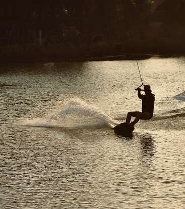A person is wakeboarding on a calm lake during sunset, creating a splash of water while holding onto a cable, with trees in the background.