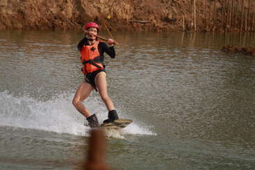A person wearing an orange life jacket and red helmet is wakeboarding on a river, holding a rope, creating splashes, with muddy banks in the background.