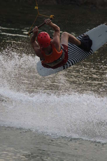 A person wearing a red helmet and orange life vest is wakeboarding, performing an aerial trick over water, creating a dynamic splash and action-packed scene.