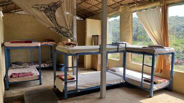 This image shows a rustic dormitory with metal bunk beds, simple bedding, open windows, natural light, earthy walls, and scenic greenery visible outside the curtains.
