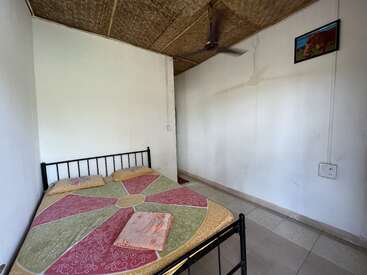 This simple room features a bed with a colorful bedspread, two towels, white walls, a ceiling fan, woven ceiling, tiled floor, and a small picture.