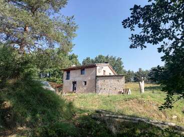 Une maison rustique en pierre se dresse sur une colline verdoyante, entourée de grands arbres et de végétation, sous un ciel bleu limpide, dégageant tout le charme paisible de la campagne et une atmosphère de tranquillité.