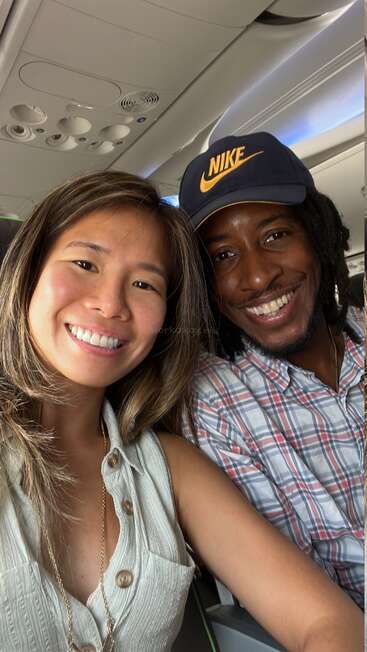 Das Bild zeigt ein Selfie von einem Mann und einer Frau in einem Flugzeug, die beide in die Kamera lächeln, wobei der Mann eine Nike-Mütze trägt.