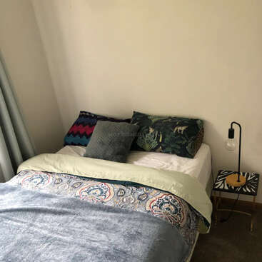 The image depicts a cozy bedroom with a neatly made bed, featuring a white comforter, patterned blanket, and pillows, accompanied by a small table with a lamp beside it.
