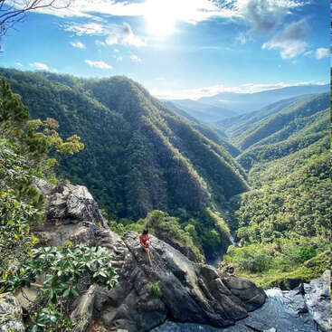 Das Bild zeigt eine Frau, die an einem sonnigen Tag auf einem Felsen mit Blick auf ein üppiges, grünes Tal sitzt, durch das ein Fluss fließt.