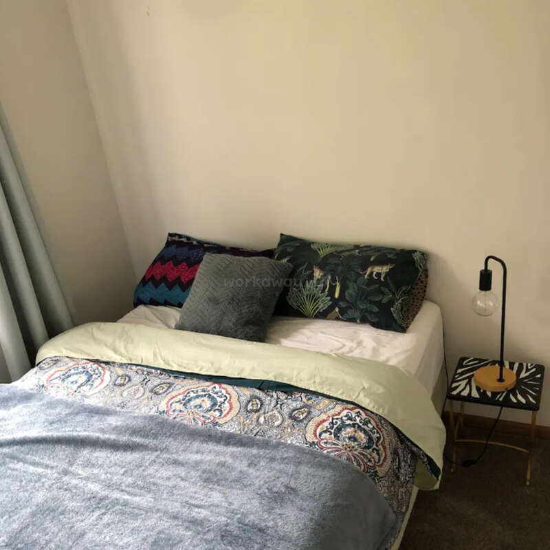 Das Bild zeigt ein gemütliches Schlafzimmer mit einem ordentlich gemachten Bett, einer weißen Bettdecke, einer gemusterten Decke und Kissen sowie einem kleinen Tisch mit einer Lampe daneben.