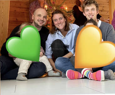 The image depicts a family of four sitting on the floor in front of a Christmas tree, with two large hearts superimposed over the photo, one green and one yellow.