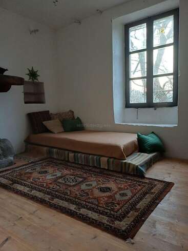 Una habitación acogedora y minimalista con un colchón en el suelo, una manta de rayas de colores, cojines verdes, una alfombra estampada, un gran ventanal, paredes blancas, suelo de madera y una pequeña planta.