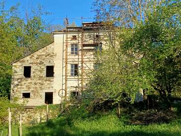 Una antigua casa de piedra se alza parcialmente oculta entre los árboles y la hierba, con andamios en la fachada, lo que sugiere que se están llevando a cabo obras de renovación o construcción en un entorno rural.