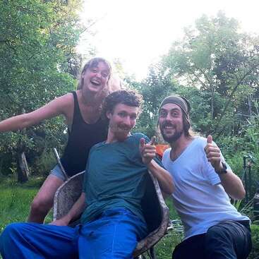 Das Bild zeigt drei Personen, die für ein Foto in einem üppigen Garten posieren, umgeben von Bäumen und Pflanzen, und vermittelt ein Gefühl von Freude und Kameradschaft.