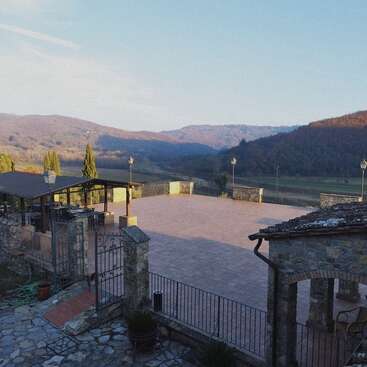 Esta imagen muestra una amplia terraza de piedra con barandilla metálica, rodeada de colinas campestres, árboles, edificios rústicos de piedra y una zona cubierta para sentarse bajo un cielo azul.