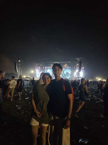 Una pareja sonriente aparece en primer plano en un concierto nocturno al aire libre, con un gran escenario iluminado en el que se lee "VASCO" y una multitud de personas detrás.