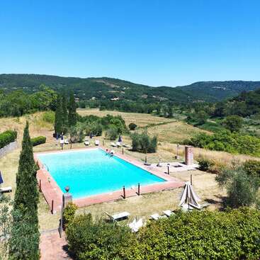 Una serena piscina al aire libre rodeada de tumbonas y sombrillas, situada en medio de hermosas colinas verdes, campos y árboles bajo un cielo azul despejado.