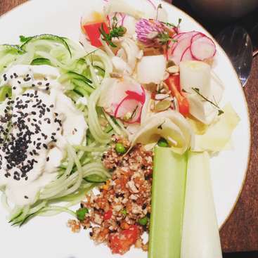 L'image représente une assiette blanche à bord doré, contenant une salade de divers légumes, dont du concombre, du céleri et du radis, accompagnée d'un assortiment de graines et de semences.