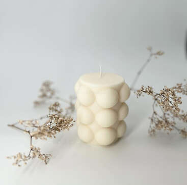 Une bougie blanche à la texture unique, avec des bulles circulaires en relief, est posée sur une surface blanche, entourée de délicates fleurs séchées, créant une esthétique douce, minimaliste et élégante.