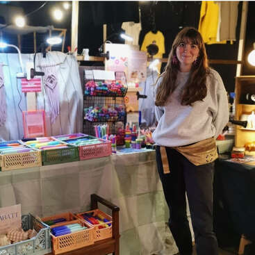Une femme sourit à un étal de marché coloré, présentant de l'artisanat, des bougies et des objets dynamiques sous des lumières chaudes, créant ainsi une atmosphère accueillante et créative.