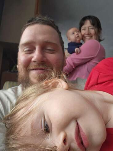 A cheerful family selfie with a bearded man smiling, a playful child in front, and a woman holding a baby in the background, all happy together.