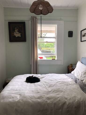 Um quarto aconchegante com uma cama bem arrumada, um gato preto enrolado, obras de arte nas paredes, uma janela com cortina e um abajur de vime.