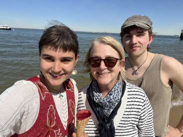 Três pessoas sorrindo na praia num dia ensolarado. Uma delas segura um sorvete. O mar e um barco aparecem ao fundo.