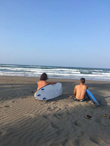 Dos hombres sin camiseta están sentados en una playa de arena, cada uno con una tabla de surf en la mano, mirando al océano, con un cielo azul despejado encima.