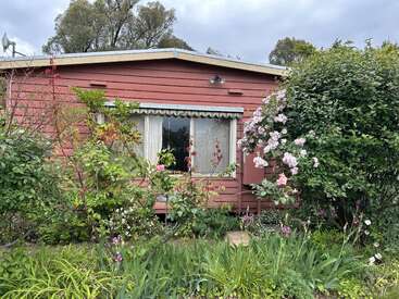 Une maison rouge chaleureuse aux fenêtres bordées de blanc est entourée de plantes verdoyantes, de fleurs roses épanouies et d'un charmant jardin bien entretenu par une journée nuageuse.