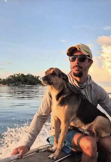Ein Mann mit Mütze und Sonnenbrille sitzt in einem Boot mit einem Hund auf dem Schoß, umgeben von Wasser, unter einem klaren, sonnigen Himmel.