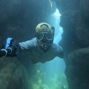 A snorkeler with a yellow cap and snorkel mask swims through a narrow underwater canyon, surrounded by rocky walls, capturing the moment with a selfie stick.