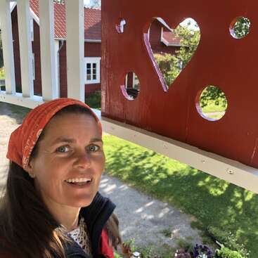 L'image représente une femme debout sous un porche, portant un foulard et une veste rouges, avec derrière elle un mur rouge orné de découpes en forme de cœur.