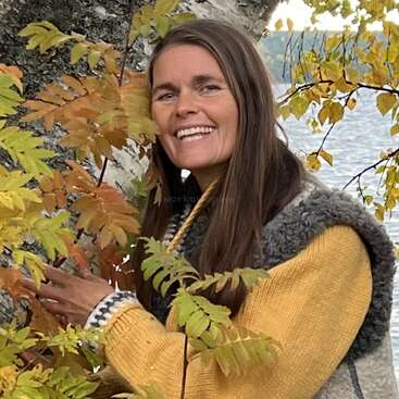 Eine lächelnde Frau mit langen braunen Haaren steht an einem Baum mit Herbstblättern, trägt einen gelben Pullover und eine graue Weste, mit Wasser und Laub im Hintergrund.
