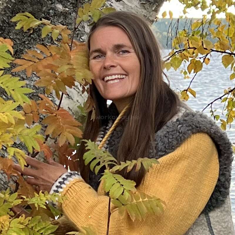 Une femme souriante aux longs cheveux bruns se tient près d'un arbre aux feuilles d'automne, vêtue d'un pull jaune et d'un gilet gris, avec de l'eau et du feuillage en arrière-plan.