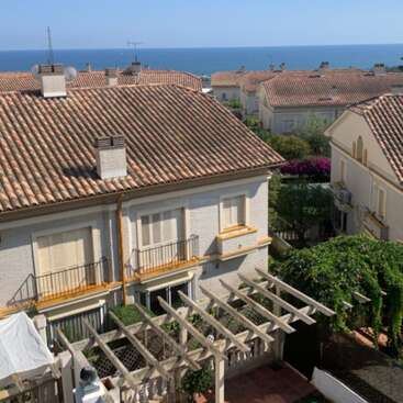 La imagen representa una serena ciudad de estilo mediterráneo, con una casa blanca con tejado de terracota, balcón y pérgola, sobre el fondo de un mar tranquilo.