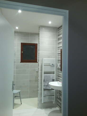 This image shows a modern bathroom with grey tiled walls, a small window, white towel radiator, wall-mounted sink, a grey towel, and a chair in the shower area.