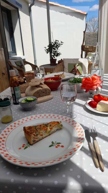 A sunlit table set for lunch with quiche, bread, fresh vegetables, water, salad, and a red rose in a vase, outdoors on a patio.
