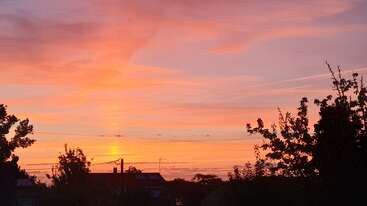 A vibrant sunset fills the sky with warm orange, pink, and purple hues. Silhouetted trees and rooftops create a peaceful, serene evening atmosphere below.