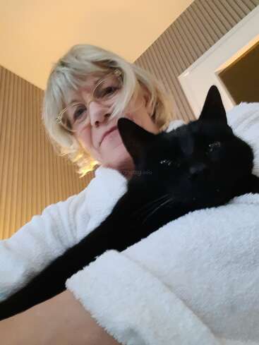A woman with blonde hair and glasses, wearing a white robe, cuddles a relaxed black cat in a cozy, warmly-lit room with striped wallpaper.