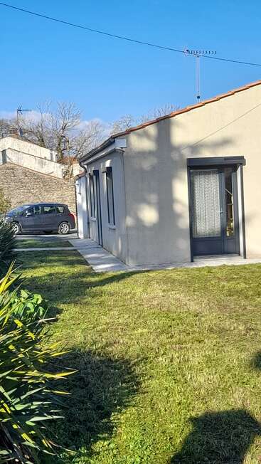 A single-story house with cream walls and large windows, surrounded by a green lawn and plants. A car is parked beside a stone wall.