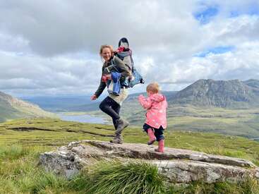 A woman hikes through grassy hills carrying a baby on her back, while a small child in pink boots and raincoat walks beside her, scenic mountains behind.