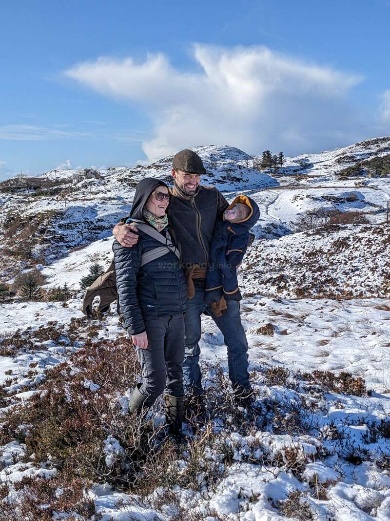 The image depicts a family of three standing in a snowy landscape, with the man holding a baby and a woman wearing a backpack.