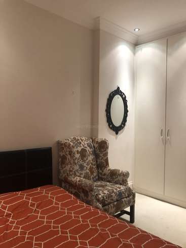 Das Bild zeigt ein Schlafzimmer mit einem Bett, einem geblümten Sessel und einem Kleiderschrank, mit weißen Wänden, einer rot gemusterten Bettdecke und einem schwarzen Spiegel an der Wand.